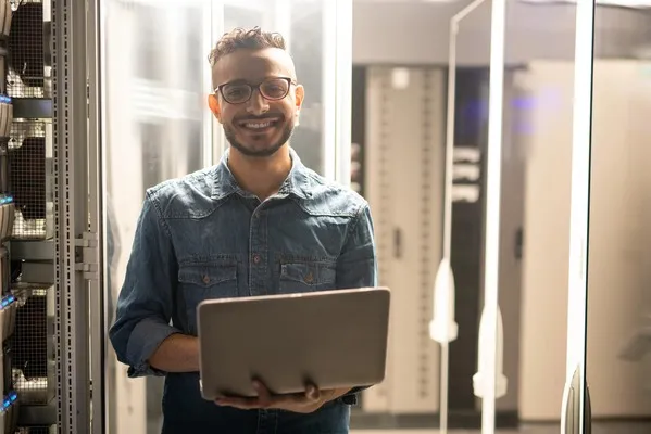 Hombre con una laptop sonriendo frente a servidor web, es una imágen ilustrativa de que es webhost